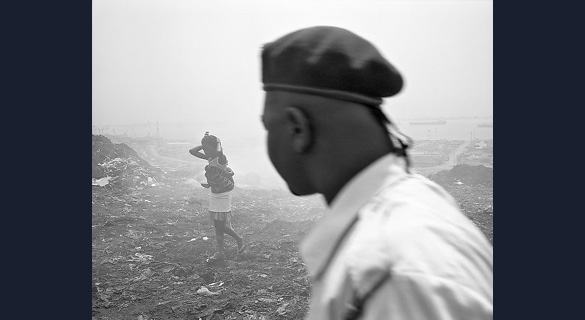 WOMAN AND HER BABY, ROQUE SANTEIRO MARKET: Conflict between Luanda’s population and its governing elites forms an undercurrent to this photograph of a young woman carrying a baby across litter-strewn ground, observed by a man wearing a military beret.