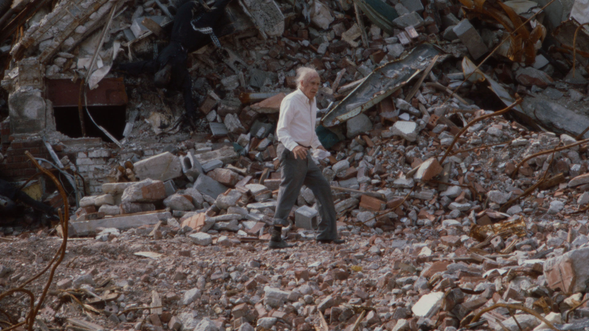 An elderly man walks in front of a pile of rubble