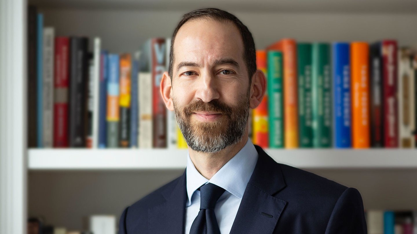 A white man with a short beard in a suit and tie, standing in front of a bookshelf