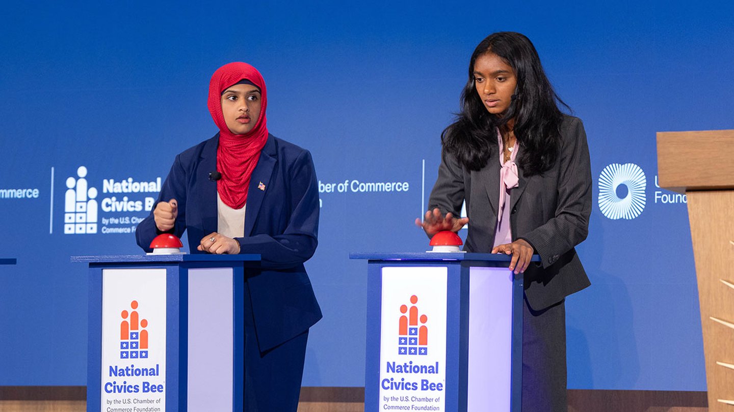 Two women stand at podiums with their hands over buzzers at the National Civics Bee